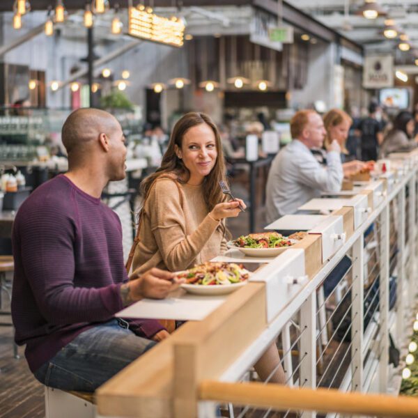 A man and woman enjoy a meal inside the Anaheim Packing House. They are sat at a white railing-like table with aesthetic lights and greenery surrounding them.