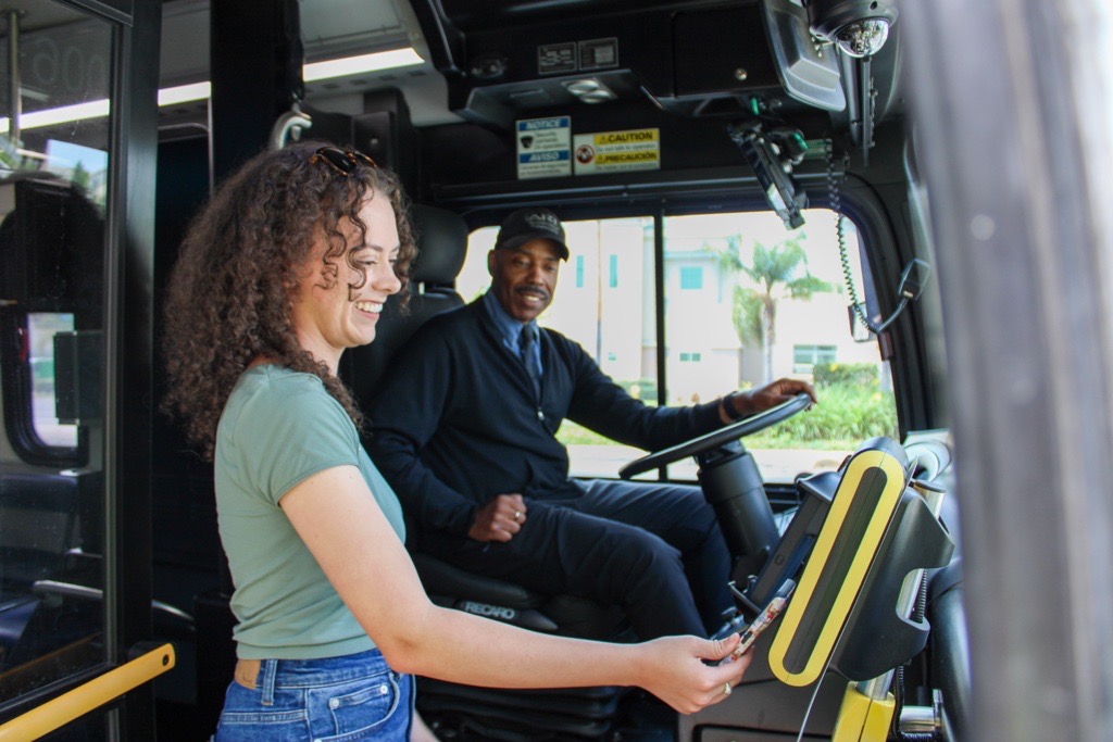 A woman entering a bus is holding her phone out to use the contactless payment option. The bus driver is smiling while holding the steering wheel.