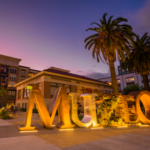 The Muzeo building at dusk with a large art instillation of letters spelling out 'MUZEO'. A purple and pink sunset light up the background behind the building. .